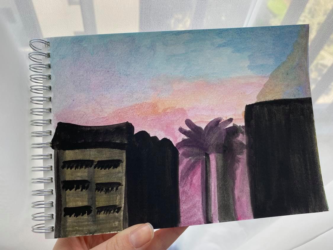Painting in a sketchbook showing the rough outlines of buildings and palm tree against a sunset background
